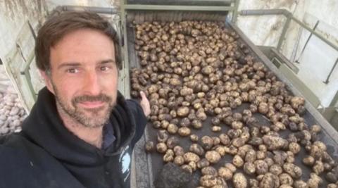 A man in a black hoodie stands in front of a conveyor belt covered in potatoes