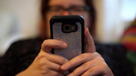 A woman holds out a mobile phone out towards the camera with both hands. Her face is blurred but she is wearing glasses and has dark hair.