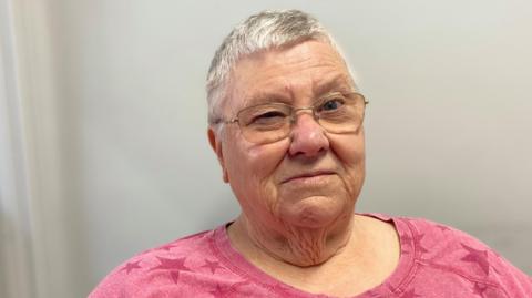 A woman with short grey hair and glasses is looking at the camera. She is wearing a pink top with a star pattern on it.