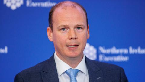Gordon Lyons. He is mostly bald with a bit of short dark hair and blue eyes. He is wearing a dark blue blazer with white shirt and light blue tie. A blue background is behind him with NI Executive branding on it.
