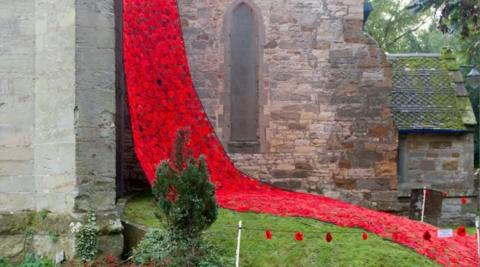 Hundreds of red knitted poppies hang down the side of a grey stone and brick building with a narrow archway in the middle. Some poppies are also covering the grass.