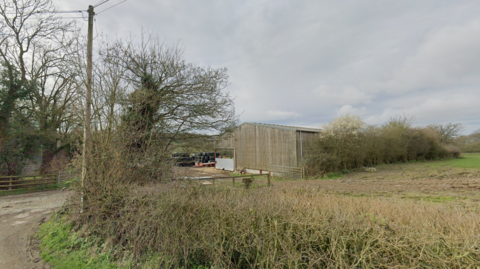 A track leads off a road to a area with a barn, surrounded by trees. A field is next door.