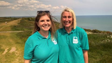 On the left Anna has long brown hair and wears a pair of sunglasses on her head. On the right, Susan's hair is a blond bob. Both are wearing turquoise polo shirts with the Beachy Head Ambassadors logo. They are smiling enthusiastically. Behind the women you can see the footpath along the cliff edge at Beachy Head and the view out to sea.  