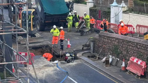 A sinkhole in a road, surrounded by equipment and workers wearing high-visibility clothing. There is scaffolding on buildings on either side of the road affected by the sinkhole, and a work truck in the vicinity.