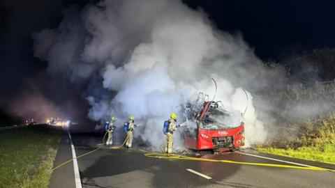 Smoke fills the air as firefighters battle a fire on a red bus.