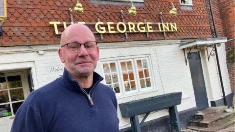 A bald man standing outside a pub. There is a gold sign on the pub which reads "THE GEORGE INN".