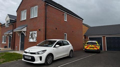Two police vehicles are parked on the drive of a newbuild house. One is a white car without any police markings on. A police officer is sat in the driver's seat. The other car has full police colours. There is a silver foil sheet over the downstairs window of the house.