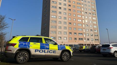 A police car parked in front of a high rise tower block in the evening sun