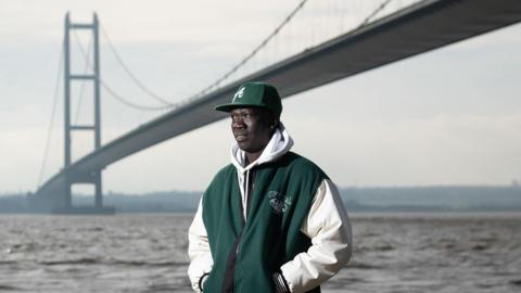 Anei smiles as he looks ahead, with the Humber Bridge in the background. Anei is wearing a bottle green cap and matching hooded jacket, blue jeans and white trainers.