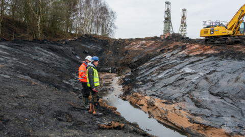 Two people in hard hats and high vis vests stood next to a yellow digger working at a mud filled site with Clipstone Colliery headstocks in the background