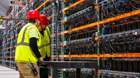 Two technicians wearing PPE walking past racks of equipment in a data center for cryptocurrency mining, cloud services and AI computing in a large warehouse