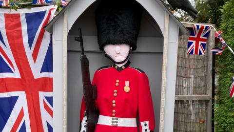 A scarecrow of a King's Guard. It is dressed in a red jacket, a tall black fluffy hat and is stood with a replica gun. There is a Union Jack and bunting hanging next to it.