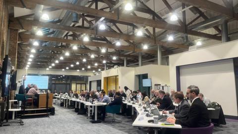 A council meeting room- with councillors sitting in rows on white tables.