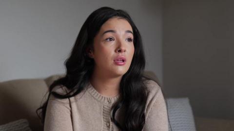A woman with dark hair and beige cardigan. She is sitting on a couch.