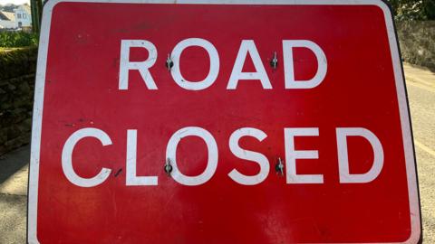 A Road Closed sign with white writing and a red background, a white border, zoomed in so you can only see parts of the road at the side of the sign.