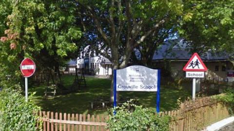 A picture of the front school yard of Grouville School. There are many trees and the building is visible in the background. The school's name sign is visible in the foreground. 