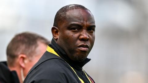 A headshot of Benni McCarthy looking over his right shoulder, wearing a black top with a yellow lanyard around his neck.