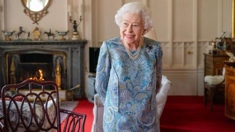 Queen Elizabeth II is standing inside a room Windsor Castle on April 28, 2022 wearing a blue dress and pearls. She is smiling