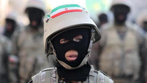 A young person wearing a balaclava, fatigues and a helmet sporting an Iranian flag looks off camera, with blurred military figures in the background.