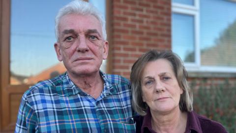 A man and a woman are standing next to each other outside a house. The man is wearing a checked top and sunglasses are in his pocket.  The woman is wearing a purple top. 