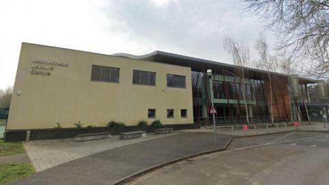 A large pale yellow building with windows adjoins a glass fronted building 