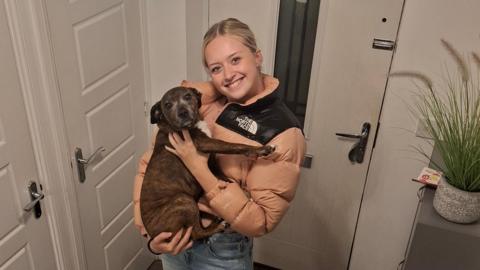 A young woman is holding a dog while smiling at the camera in a house, infront of the front door.