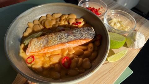Crispy salmon fillet in a big grey bowl on top of a bed of creamy butterbeans, presented on a wooden tray with bowls of garlic and chilli.