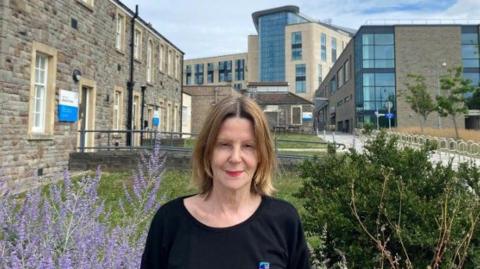 Maria Kane is standing in a garden with Southmead Hospital's main building behind her. She has dark blonde shoulder-length hair and is wearing a black top.