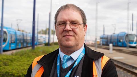 Chris Bruce standing at a tram depot. Trams and tracks can be seen behind him. He is wearing an orange high-vis vest and has a blue lanyard on. 