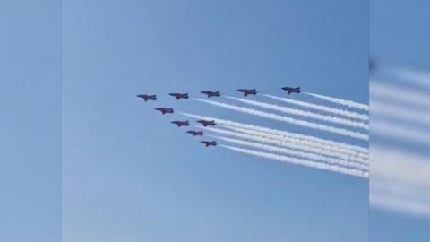 Red Arrows aircraft flies in a ten formation with white smoke trail out the back, blue sky and crowds looking from the ground.