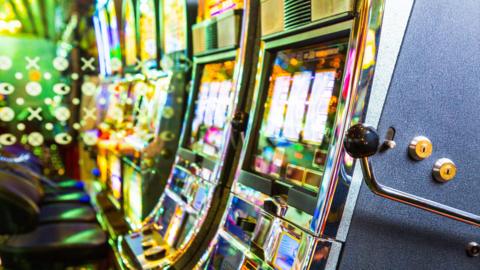 A close-up of a row of brightly lit slot machines. The machines are sitting side‑by‑side in a tight line, their reflective chrome edges catching the ambient neon.