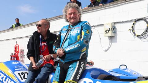 John, who is wearing his leathers, sits on his blue and yellow sidecar, smiling, next to another man.
