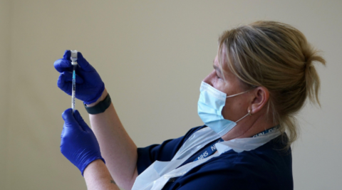 A blonde nurse with protective coverings on her face, hands and chest holds up a needle and vaccine vial.