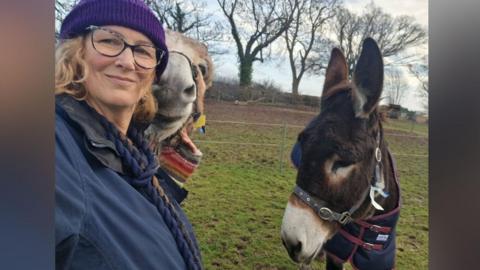 A woman takes a selfie with two donkeys in a field. The blonde woman is wearing glasses, a purple beanie and navy jacket. The donkeys are to her right and a railing, hedges and trees beyond.