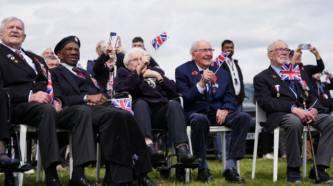 A group of veterans (from left to right, two men, a woman and two other men) sitting on white plastic chairs holding Union flags while watching a fly past in a field