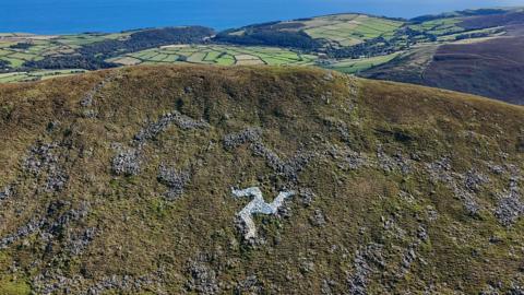 A fairly close view of the grassy hillside with a ridge that stretches from left to right, with a mass of green fields below in the distance and the sea beyond. Below the top of the ridge of North Barrule is a white Three Legs of Man symbol, clearly visible on a sunny day.