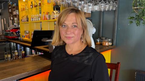 A woman with short blonde hair and a fringe who is wearing a black blouse. She is standing in front of the bar and looking at the camera.
