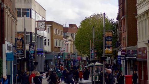 Wolverhampton city centre. There are large crowds of people walking along outside shops. There is a Costa coffee shop and a number of bright signs in view. 