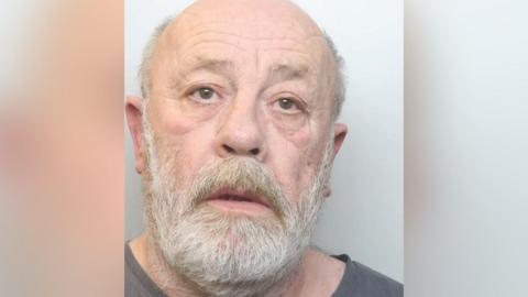 A mugshot of a bald man with a white beard and wearing a dark grey top.