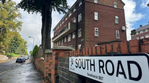 A picture of the South Road road sign, reading South Road, Sandwell, Smethwick B67. Cars are parked on the right hand side of the road, and a block of flats is visible to the right.