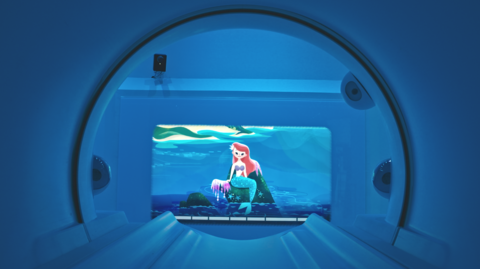 A view from inside an MRI scanner, looking through an archway with a cartoon image of a mermaid with red hair and a blue tail sitting on a rock in the sea at the end of the room. The room is lit in blue light.