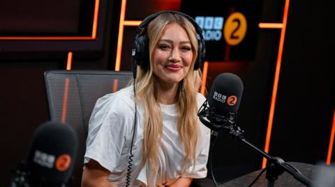 Hilary Duff in the Radio 2 studio. She has blonde hair and wears a white T-shirt