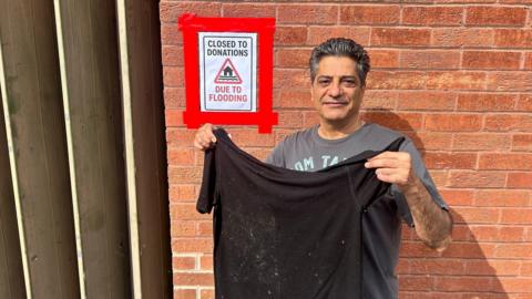 A man stands in front of a wall holding a black t-shirt which has marks on it. There is a sign behind him which reads 'Closed to donations: Due to flooding".