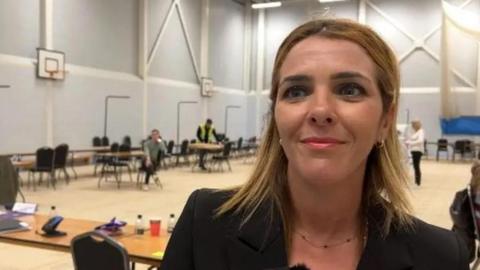 A woman with brown/blonde hair is on the right of the photo wearing a dark jacket. She is standing in a sports hall.