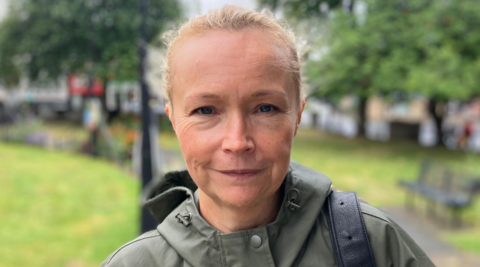 Helen is looking directly at the camera and smiling in this tightly-cropped image. She has blonde hair tied back and is wearing a green mac/waterproof with a bag over her shoulder.