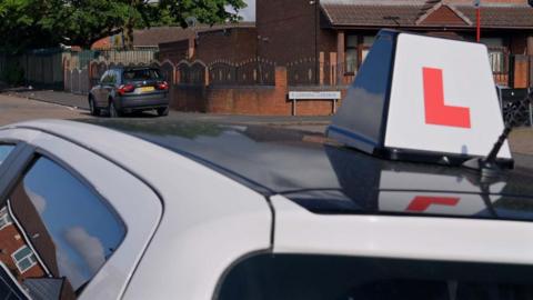 An L plate on the top of a car on a road with houses beyond.