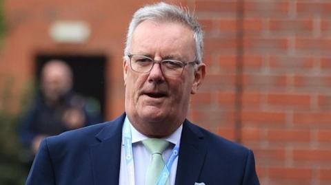 Mike Nesbitt with grey hair and glasses, wearing a dark navy suit jacket. light coloured shirt and pale green tie