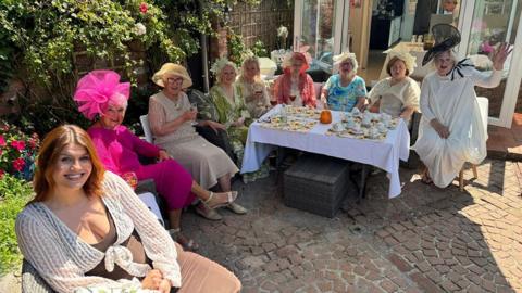 Paula Gamester with the other ladies, all dress in their finery, sitting round a table in the garden smiling at the camera