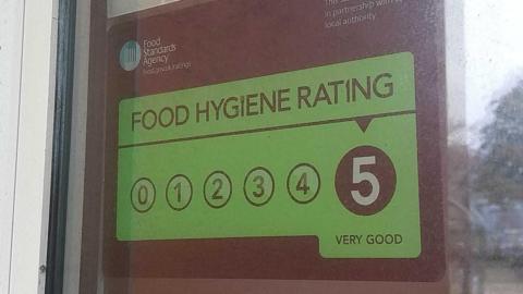 A green food hygiene rating sticker on displayed in the window of an eatery. The sticker displays that the eatery in question has a 'very good' rating of five - the highest possible. The logo for the Food Standards Agency is alos visible on the sticker.