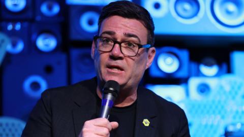 Andy Burnham speaks into a microphone at an event. He is in front of a blue wall and wears a dark suit and top.